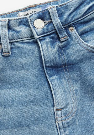 Lysblå denimjeans med brede ben, der har en høj talje, synlige syninger og et mærkelogo ved taljen. Sølvfarvet knaplukning.