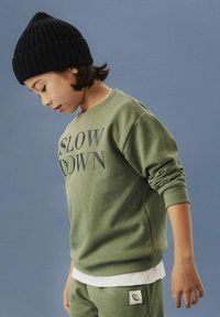 Niño con un gorro de punto negro y sudadera verde que dice "REDUCE LA VELOCIDAD", mirando hacia abajo sobre un fondo azul liso.