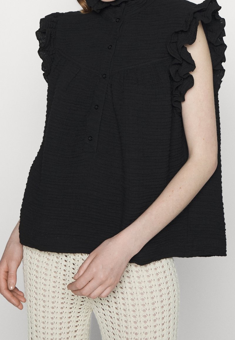 Femme portant un chemisier noir sans manches avec des épaules à volants et des boutons sur le devant, associé à un pantalon blanc en crochet.