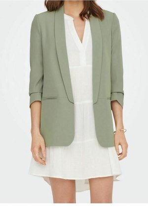 Kobieta w jasnozielonej marynarce na białej sukience z dekoltem w serek, z podwiniętymi rękawami i minimalną biżuterią na dłoniach i nadgarstkach.