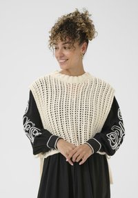 Jersey de color crema de punto con textura de tejido abierto, superpuesto a una blusa negra de manga larga que presenta bordados decorativos blancos en las mangas.