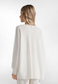 Pull en tricot crème surdimensionné avec une finition texturée, des manches larges et une petite étiquette rose à l'arrière du col. Coupe douce et décontractée.