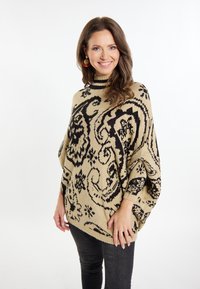 Maglione oversize lavorato a maglia di colore beige con motivi astratti neri, colletto alto e maniche larghe. Abbinato a jeans scuri.