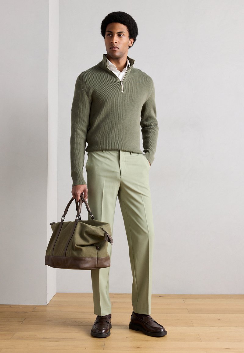 Grøn ribstrikket sweater med lynlås i kraven, lysegrøn skræddersyet buks, brune lædersko og en grøn canvas taske med brune detaljer.