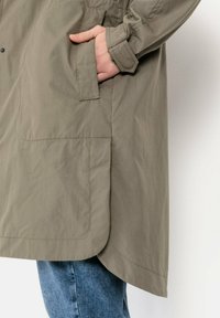 Olivgrüne leichte Jacke mit sichtbarem Nähten und einer Seitentasche. Der Saum ist abgerundet und der Stoff wirkt glatt mit einer matten Oberfläche.