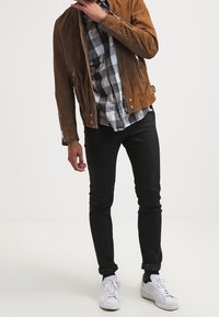 Giacca in camoscio marrone con cerniere, indossata sopra una camicia a scacchi bianca e nera. Pantaloni neri aderenti e sneaker bianche completano l'outfit.