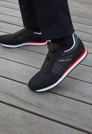 Mustat ja punaiset Armani Exchange -lenkkarit, päällä mustat housut puulattialla, fokus sivun logossa ja pohjan yksityiskohdissa.