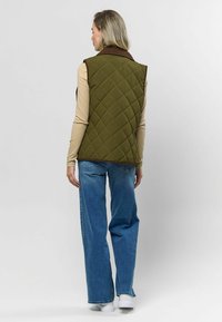 Oliwkowa pikowana kamizelka z brązowym kołnierzem, noszona na dopasowanej bluzce z długim rękawem, w zestawie z niebieskimi spodniami o szerokich nogawkach i białymi trampkami.