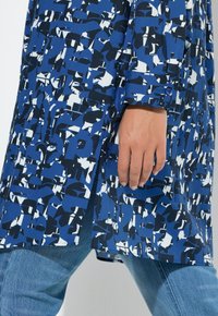 Blauw shirt met abstract zwart-wit patroon, lange mouwen, met knopen afgewerkte manchetten, getoond naast een hand. Combineer met lichtblauwe spijkerbroek.