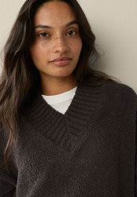 Tumepruun tekstuuriga kampsun laia V-kaelusega ja soonikkoega, kantud valge pikakarvalise särgi kohal. Sile ja mugav materjal.