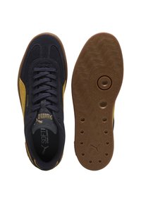 Baskets Puma noires et moutarde avec lacets, semelle intérieure douce visible, et semelle en caoutchouc marron présentant des motifs circulaires et le logo Puma.