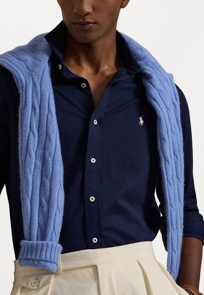 Ναυτικό μπλε πουκάμισο με άσπρα κουμπιά και λογότυπο, καλυμμένο με έναν ανοιχτό μπλε πλεγμένο πουλόβερ ριγέ στους ώμους, σε συνδυασμό με κρέμ παντελόνια.