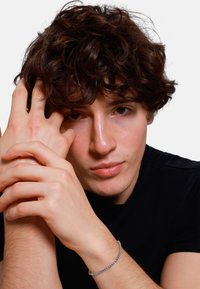 Un giovane con capelli ricci e castani posa con le mani vicino al viso, indossando un sottile braccialetto d'argento su un polso nudo, su uno sfondo bianco.