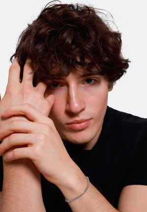 Un giovane con capelli ricci e castani posa con le mani vicino al viso, indossando un sottile braccialetto d'argento su un polso nudo, su uno sfondo bianco.