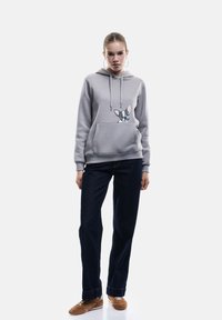 Felpa grigia chiara con grafica di un bulldog francese sulla tasca, abbinata a jeans blu scuro e sneakers marroni. Finitura in tessuto liscia.