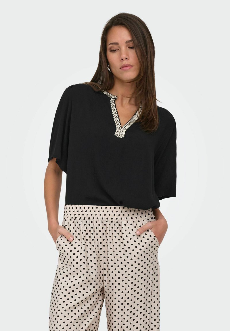 Femme portant un chemisier noir avec une broderie blanche de feuilles sur le col et un pantalon beige à pois noirs, mains dans les poches, regardant vers le bas.