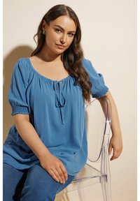 Blå, lättviktig topp med rundad halsringning, knytdetalj och elastiska puffärmar. Bärs med jeans, sittande på en genomskinlig stol.