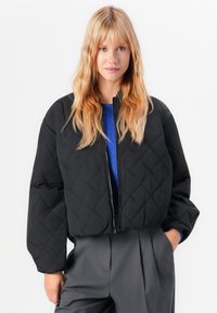 Femme blonde portant une veste zippée matelassée noire, un pull bleu et un pantalon plissé gris, se tenant avec une main dans la poche.