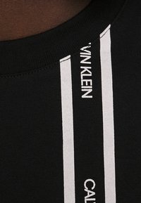 Černá mikina s kulatým výstřihem, zdobená bílými svislými pruhy a logem "CALVIN KLEIN" na levé straně. Měkký textilní materiál.