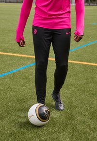 Rosa langärmliges Sportoberteil, schwarze enge Hose mit Logo, schwarzer und goldener Fußball auf einem Kunstrasenfeld.