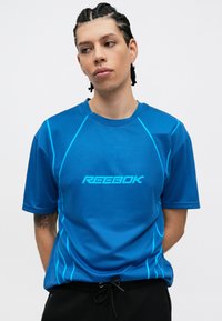 T-shirt atlético azul com mangas curtas, apresentando riscas em teal e o logótipo "REEBOK" no peito. Material suave e leve.