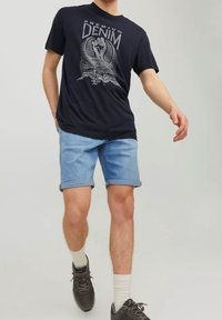 Man wearing navy graphic "Premium Denim" t-shirt, light blue rolled-up denim shorts, white socks, and black sneakers walking forward.