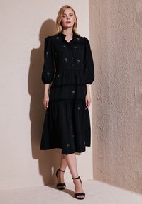 Rochie midi neagră, cu fuste în straturi, mâneci bufante, nasturi în față și accente de broderie florală albastră. Material neted cu o croială lejeră.