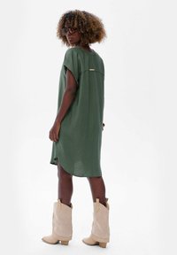Oliwkowa sukienka do kolan z krótkimi rękawami i zaokrąglonym dołem. Posiada detal z tyłu i jest zestawiona z beżowymi kozakami do połowy łydki.