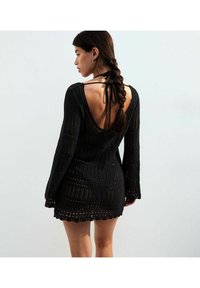 Abito nero in maglia con maniche lunghe, caratterizzato da una profonda scollatura a V sul retro, intricate lavorazioni a punto e orlo scallop. I lacci al collo completano il design.