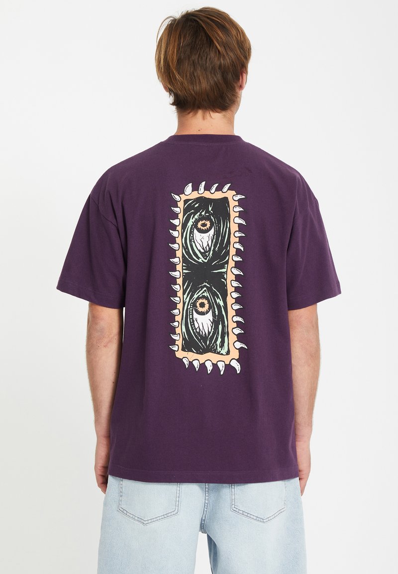 Paarse katoenen T-shirt met een grote grafische afbeelding op de rug, bestaande uit twee gestileerde ogen en tandachtige accenten rond de rand. Korte mouwen.