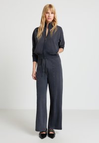 Blonde Frau steht, trägt dunkelgraue Jacke mit Reißverschluss, passende weit geschnittene Hosen und schwarze spitze Schuhe mit Riemen und Nieten.