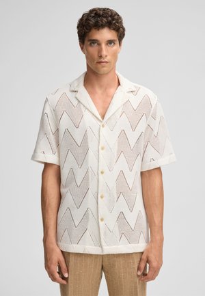 Camicia bianca a maniche corte con motivo a zigzag, realizzata in un tessuto leggero e texturizzato, caratterizzata da un colletto a punte e bottoni.