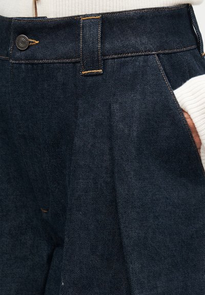 Σκούρο μπλε τζιν με κίτρινα ράμματα, κλείσιμο με κουμπιά, θηλιές για ζώνη και ένα χέρι μερικώς στην δεξιά τσέπη.