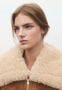 Bruine suède jas met een pluizige, crèmekleurige shearlingkraag en zilveren rits. Eenvoudig ontwerp, gladde textuur, neutraal kleurenpalet.