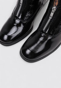 Botines de charol negro con un diseño elegante, que cuentan con una punta redondeada y detalles de cremallera de metal plateado en el lateral.