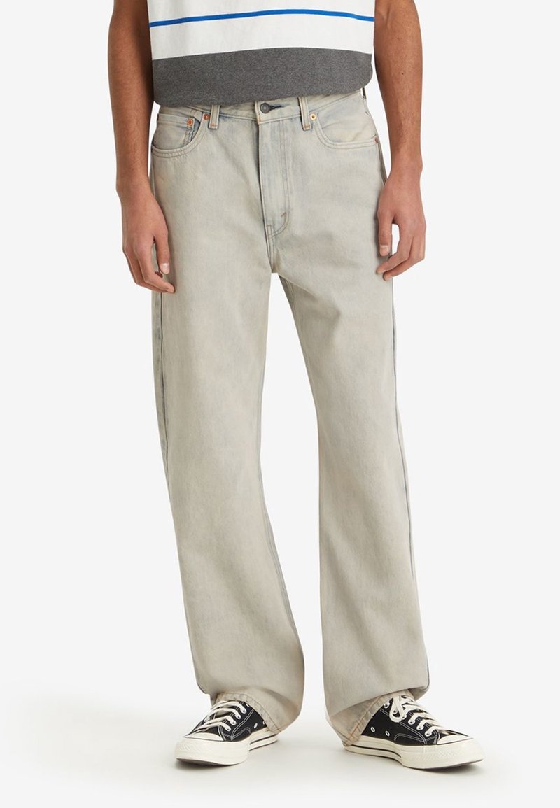 Jean en denim gris clair délavé avec une coupe décontractée, des jambes droites et des coutures orange, associé à des baskets basses noires.