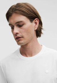 Camiseta blanca de manga corta con textura acanalada, escote redondeado y un pequeño logotipo en el pecho. Vista en primer plano.