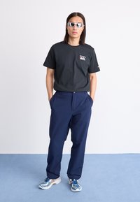 Zwarte katoenen t-shirt met logo, gecombineerd met marineblauwe broek. Model draagt witte zonnebril en blauwe sportschoenen met witte accenten.