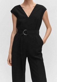 Mono negro con mangas cortas tipo cap sleeve, escote en V, cintura ceñida acentuada por una hebilla de cinturón circular y diseño de pierna ancha. Textura de tela suave.