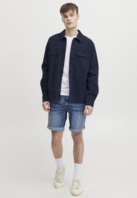 Chemise bleu marine à boutons avec deux poches poitrine, portée sur un t-shirt blanc, associée à un short en denim retroussé et des baskets blanches.