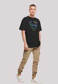 Black oversized t-shirt featuring a colorful floral Superhero logo. Paired with khaki cargo pants and white sneakers, showcasing casual style.