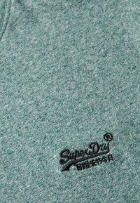 Groene gemêleerde stof met een zachte textuur heeft een geborduurd "SuperDry" logo in het zwart, gepositioneerd rechtsonder.