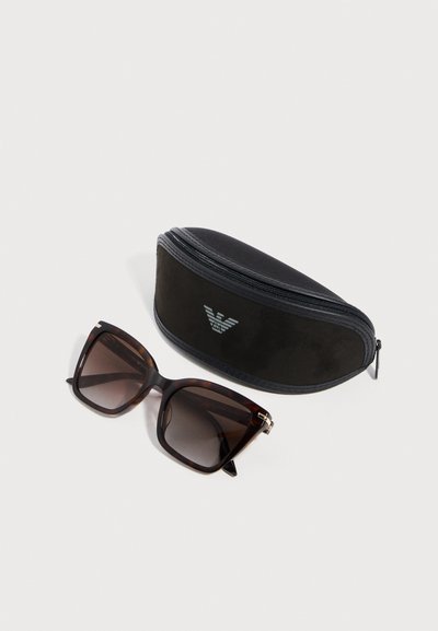 Τετράγωνα γυαλιά ηλίου με σχέδιο χελώνας και σκούρους φακούς με βαθμιαία απόχρωση δίπλα σε μια μαύρη θήκη με φερμουάρ που φέρει ασημένιο λογότυπο στο μπροστινό μέρος.