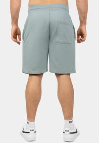 Lichtgrijze heren shorts van zacht materiaal, met een elastische tailleband, zijzakken en een achterzak met klep. Gepaard met witte sneakers.