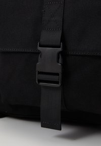 Sac messager en tissu noir avec une boucle en plastique et une sangle ajustable. Surface texturée avec des coutures renforcées pour plus de durabilité.