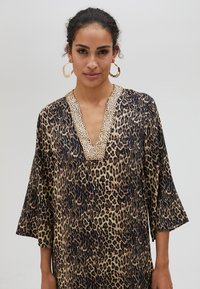 Tunika uzorkom leoparda s V-izrezom, ukrašena zlatnim rubom od perlica, s širokim rukavima i mekim, teksturiranim materijalom.