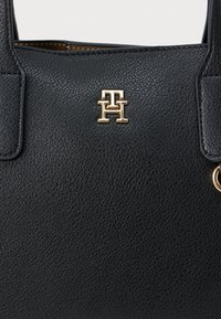 Mustast nahast käekott, millel on tekstureeritud pind, kuldse tooni logo riistvara ja kaks ülemist käepidet. Sisetükid ei ole nähtavad.