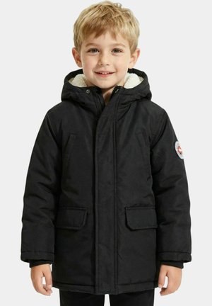 Garçon blond souriant, portant un manteau d'hiver noir à capuche avec deux poches avant et un patch rond sur la manche gauche.