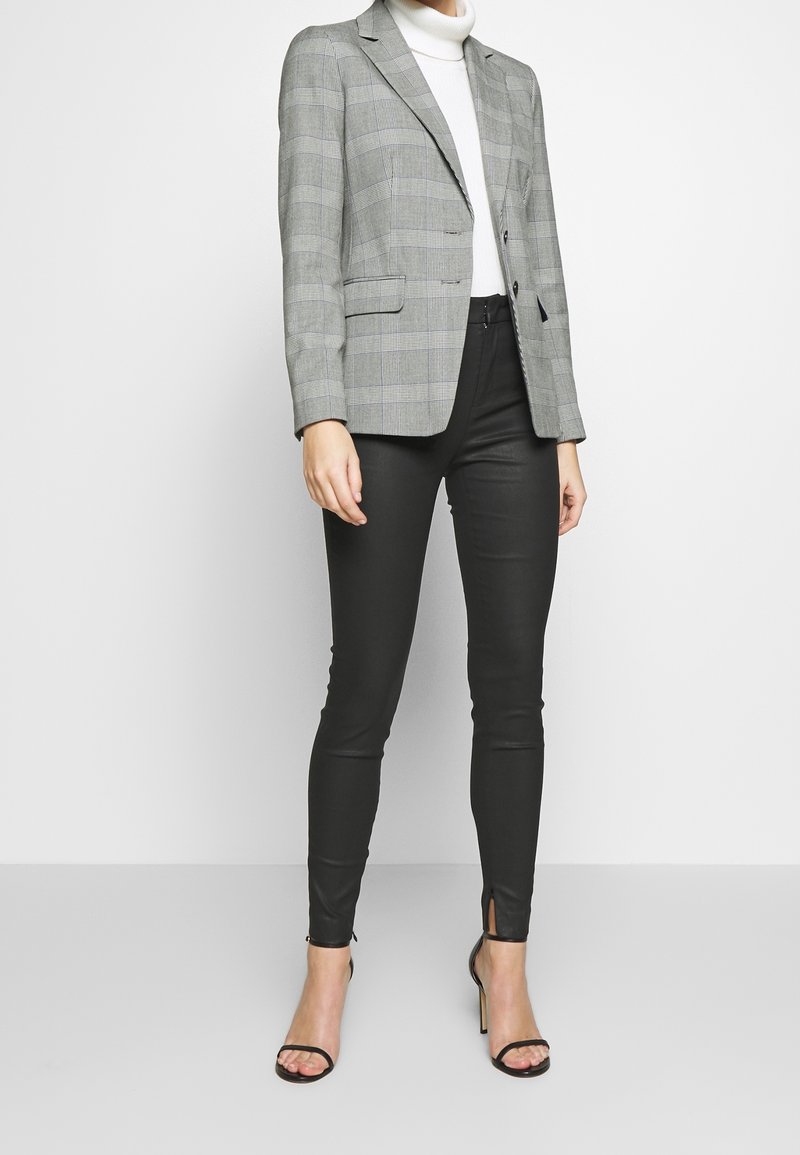 Femme portant un blazer à carreaux gris, un col roulé blanc, un pantalon slim noir avec des fentes aux chevilles et des talons hauts noirs à bouts ouverts sur fond blanc.