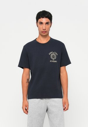 Giovane uomo con una maglietta nera "Army Corp Academy" e pantaloni grigi chiari, in piedi davanti a uno sfondo bianco uniforme.